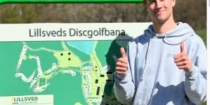 Man in a light gray hoodie stands beside a large green disc golf course map sign that reads 'Lillsveds Discgolfban,' giving a thumbs-up gesture.
