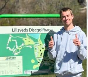 Screenshot - Lillsved Man in a light gray hoodie stands beside a large green disc golf course map sign that reads'Lillsveds Discgolfban,' giving a thumbs-up gesture.
